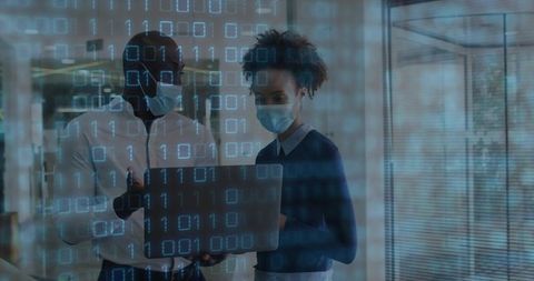 Masked colleagues analyzing cybersecurity data on laptop, collaborating in glass office now