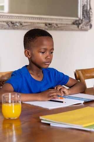 African american boy studying at dining table doing homework with orange juice