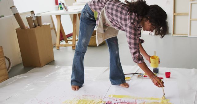 Female artist bending over and painting on a large canvas laid on the floor of a studio. She appears passionate and focused on her work. Barefoot and wearing a checkered shirt and jeans with an apron, she is using bright colors on the canvas, surrounded by painting supplies. Ideal for illustrating creative processes, studio environments, artistic expression, and contemporary art creation.