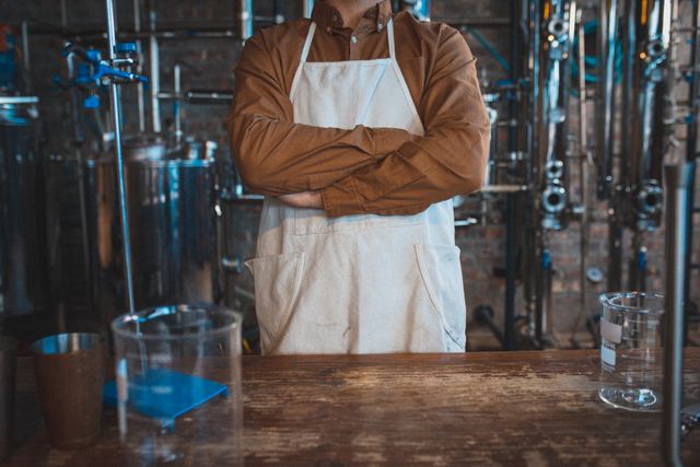 Midsection of caucasian man wearing apron at gin distillery, standing by equipment with arms crossed. work at an independent craft gin distillery business.