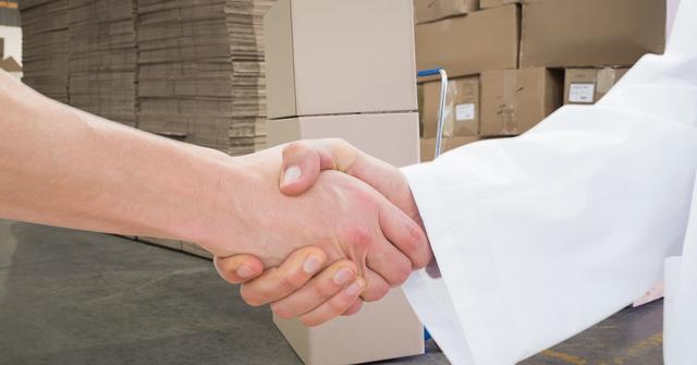 Evidently displaying two people shaking hands in a warehouse setting with stacks of boxes in the background, emphasizing successful business agreement and logistics teamwork. Ideal for illustrating concepts related to partnerships, business deals, collaboration in supply chain management, or logistics industry.