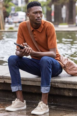 Contemplative african man sitting by urban fountain holding smartphone in rust polo