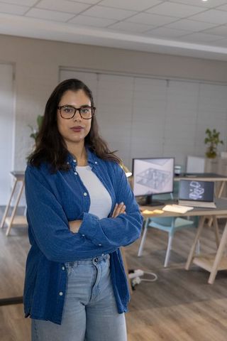 Confident designer standing with arms crossed in modern office wearing denim and glasses