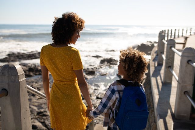 Rear view of a biracial woman wearing a yellow dress and her son enjoying time together by the sea, holding hands and smiling at each other on a promenade on a sunny day