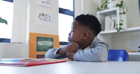 Schoolboy gazing out classroom window while sitting at desk, contemplating schoolwork