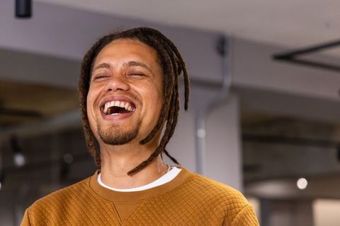 African american man laughing in modern office portrait, dreadlocks and brown sweatshirt