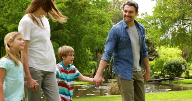 Family of four enjoying a leisurely walk in a lush green park. Parents holding hands with their children while everyone has a cheerful expression. Perfect for use in promotions centered on family values, outdoor activities, summer outings, and healthy living.