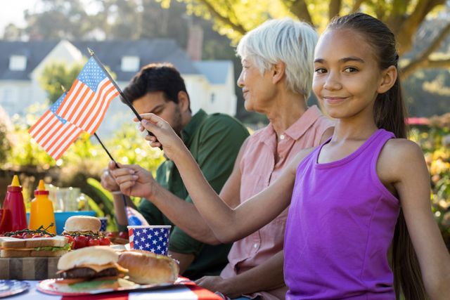 Young girl holding an American flag while sitting at a picnic table with family members. Ideal for themes of patriotism, family gatherings, summer celebrations, and outdoor activities. Perfect for advertisements, social media posts, and articles about family bonding, national holidays, and outdoor events.