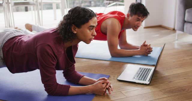Happy diverse male couple exercising, using laptop in living room. Spending quality time at home.
