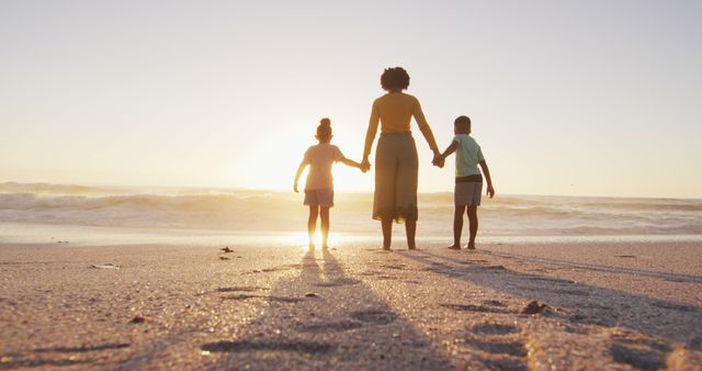 Image depicting a mother holding the hands of her two children while walking on a beach during sunset. Ideal for use in family-focused advertisements, parenting blogs, travel brochures, and lifestyle articles highlighting outdoor activities and family bonding moments.