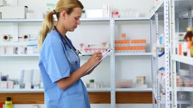 Blonde female pharmacist with stethoscope writing on clipboard while standing in a hospital pharmacy. Shelves filled with medications and pharmaceutical supplies in the background. Suitable for medical, healthcare, and professional environment concepts. Ideal for use in articles, educational content, or advertisements related to pharmacy, medicine, and healthcare professions.