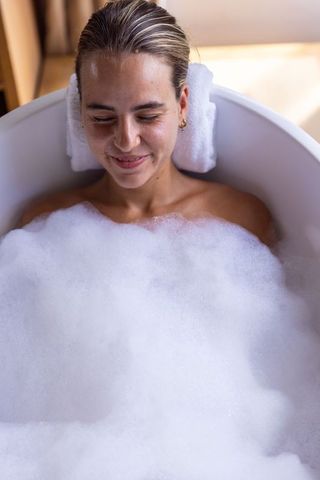 Young woman soaking in bubble bath resting head on towel wearing gold hoop earrings