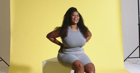 Laughing mid-adult african american woman in gray activewear sitting on block yellow backdrop
