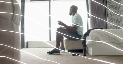 Man sitting on ottoman holding cup gazing out window in bright modern guestroom interior