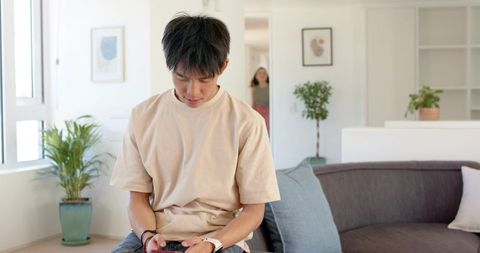 Young asian man sitting on sofa using smartphone while african woman pausing in doorway
