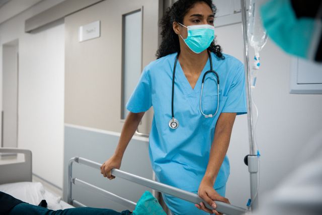 Biracial female doctor wearing face mask pushing patient in bed in hospital corridor. medical and healthcare services at hospital during covid 19 pandemic