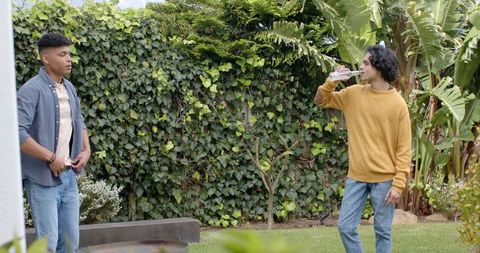 Two young men socializing in backyard garden drinking from bottle near ivy-covered fence