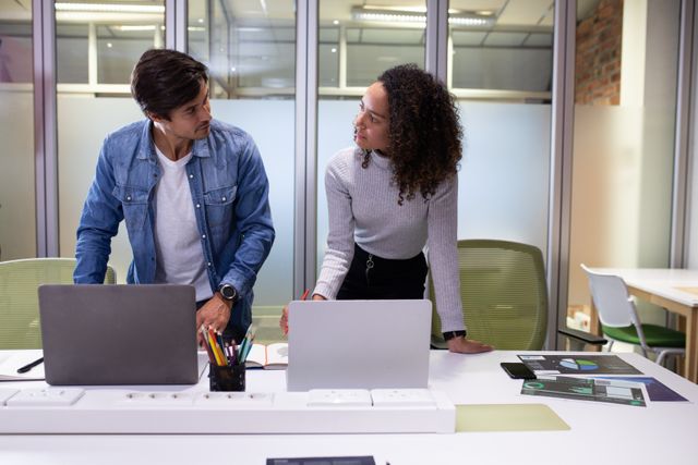 Front view of a Caucasian businessman and a biracial businesswoman working in a creative modern office together, standing by their desks with laptop computers, talking brainstorming.