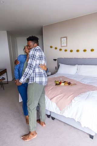 Diverse couple embracing barefoot beside cozy bed with charcuterie tray and soft blanket