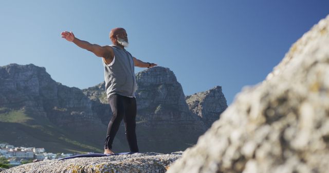 Senior man practicing yoga outdoors on a mountain top against a clear blue sky, embracing nature and fitness. Ideal for health and wellness blogs, active lifestyle promotions, and senior fitness campaigns.