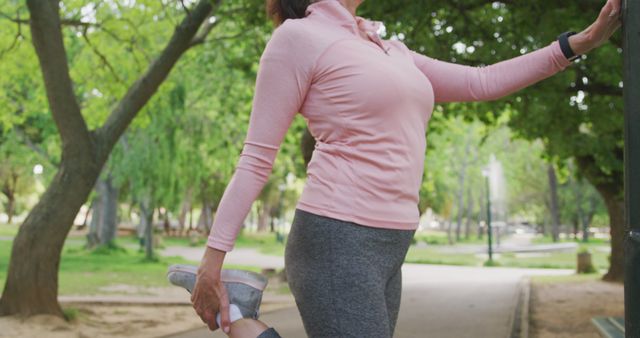 Woman in a park stretching her leg while holding onto a post, preparing for outdoor exercise or jogging. Ideal for articles or promotions related to health, fitness, women's wellness, outdoor activities, active lifestyles, exercise routines, and athletic motivation.