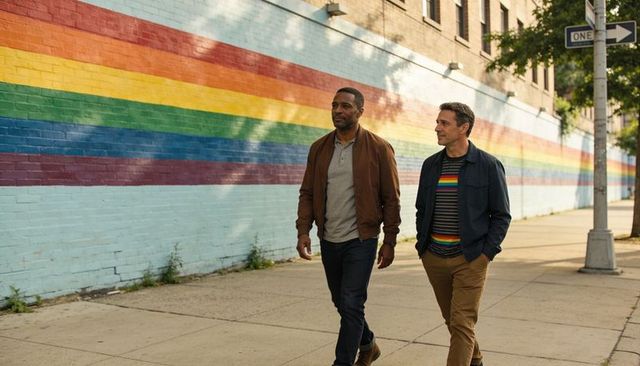 Two men walking by rainbow mural on urban sidewalk sharing relaxed conversation and smiles