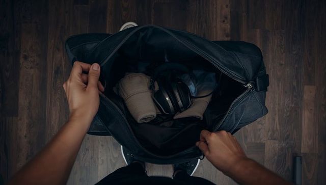 Holding open black duffel bag revealing headphones and rolled clothes on wooden floor