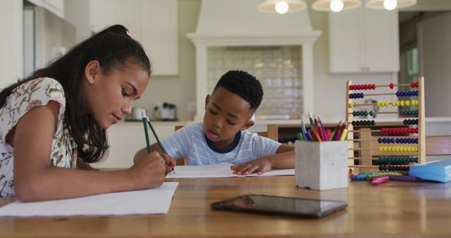Two siblings are seen concentrating on their schoolwork together at home. They are seated at a kitchen table with various study materials such as an abacus, pencils, and tablet, fostering a collaborative learning environment. This image is suitable for promoting educational activities, homeschooling, family bonding, or advertisements for educational products.
