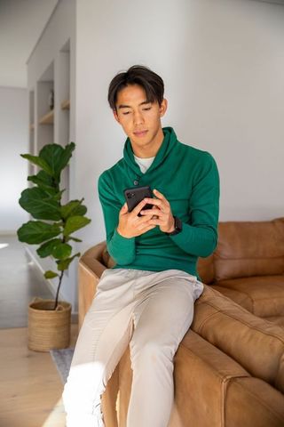 Asian man sitting on leather sofa arm scrolling smartphone in modern living room