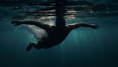 Swimmer wearing cap and goggles gliding underwater toward surface with sunbeams
