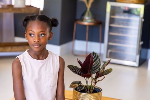 Young girl sitting in minimalist interior gazing beside gold planter with tropical foliage