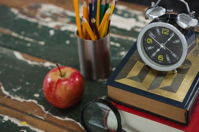 Close-up of alarm on stack of books with pen holder, apple, and magnifying glass on table