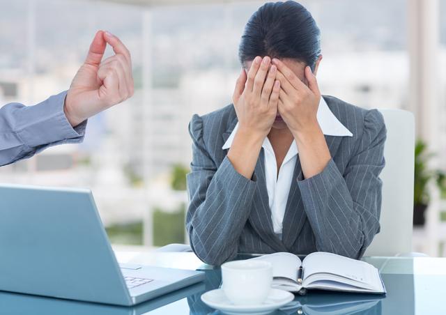 Businesswoman covering her face while being criticized by colleague's hand gesture at office desk with laptop and coffee cup. Useful for articles about workplace conflict, stress at work, professional development, and employee well-being.