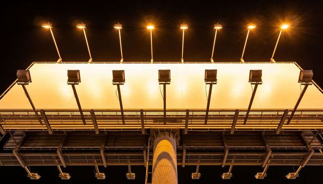 Illuminated blank billboard panel glowing above road at night showing catwalk and lamps