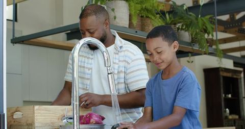 African american father and teenage son rinsing vegetables at modern kitchen sink