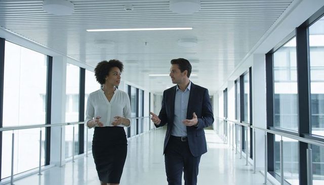 Two business professionals walking and talking in modern glass office walkway, professional discussi