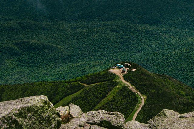 This stock photo shows an aerial view of an isolated mountain cabin surrounded by a lush green forest. The cabin is located at the top of a mountain, with a hiking trail leading up to it. The surrounding dense forest creates a tranquil and scenic atmosphere. Ideal for use in travel blogs, nature magazines, and promotional materials for tourism in remote destinations.
