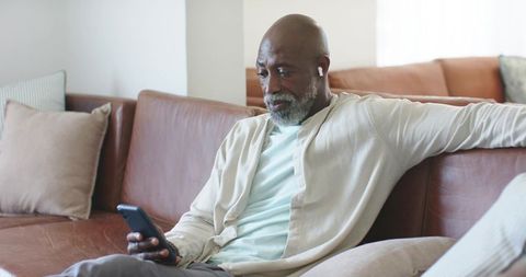 Senior african-american man sitting on sofa using smartphone and earbuds, relaxing at home
