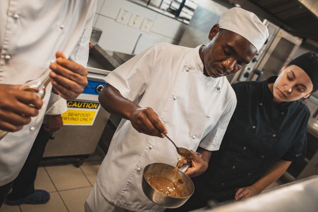 African American chef demonstrating meal preparation to a diverse group of colleagues in a busy restaurant kitchen. Ideal for use in articles or advertisements about culinary education, professional kitchen environments, teamwork in the kitchen, and chef training programs.