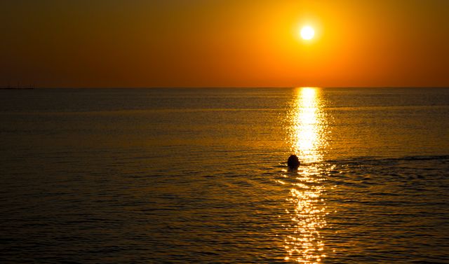 A serene scene with a person enjoying a swim during a beautiful golden sunset over calm waters. This visual is perfect for travel agencies, vacation advertisements, or inspirational content promoting relaxation and natural beauty. It can also be used in wellness blogs and nature-focused websites to convey peace and tranquility.