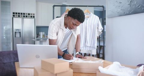 Small business owner packing clothing orders at home workstation with laptop and boxes