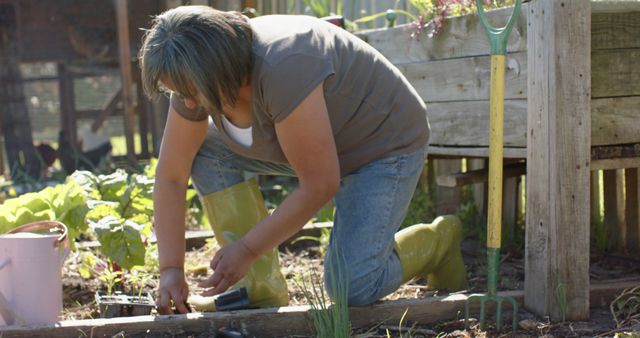 Caucasian woman kneeling and planting vegetables in a backyard garden. Wearing casual clothes and green rubber boots. Garden tools, plants, and leaves are visible. Ideal for content related to hobbies, sustainable living, horticulture, rural lifestyle, and home gardening.