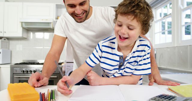 This stock photo depicts a father assisting his son with homework in a clean, bright kitchen environment. Both father and child appear joyful, enhancing the themes of family bonding and supportive parenting. This image is ideal for marketing family-oriented products, educational materials, and content related to work-life balance and parenting.