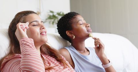 Female friends enjoying facial self-care with jade roller and gua sha on couch