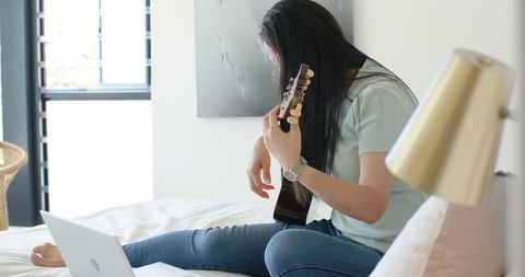Asian woman practicing acoustic guitar on bed with laptop in bright minimalist bedroom
