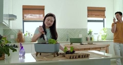 Asian couple gardening in bright modern kitchen, woman potting herb seedlings on countertop