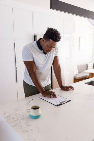 African american man leaning over bright modern kitchen island checking documents at home