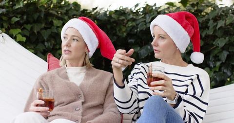 Friends wearing santa hats drinking cocktails outdoors on white lounge with ivy background