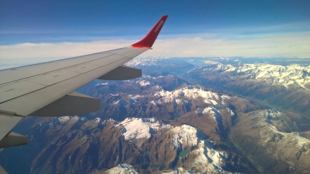 Aerial view of an airplane wing with snow-capped mountains and valleys below, bathed in clear blue sky. The image portrays the beauty of high-altitude landscapes, highlighting the contrast between the rugged mountain terrain and tranquil blue sky. Suitable for use in travel promotions, aviation industry marketing, adventure blogs, geographic or nature documentaries, and scenic view portfolios. Perfect for conveying the excitement and beauty of air travel and exploring new destinations.