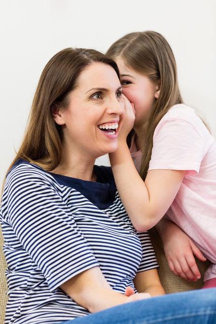 Daughter whispering a secret to her smiling mother in the living room at home. Perfect for use in family-related blogs, articles about parenting, advertisements featuring family life, and promotional materials for home and living products.
